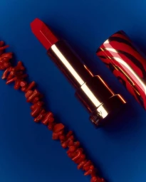 Red lipstick in a metallic case next to a coral branch on a vivid blue background by Paul Lepreux