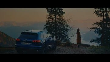 A dark blue SUV sits on a mountain top at twilight, overlooking a scenic valley. A single woman stands nearby looking at the view.