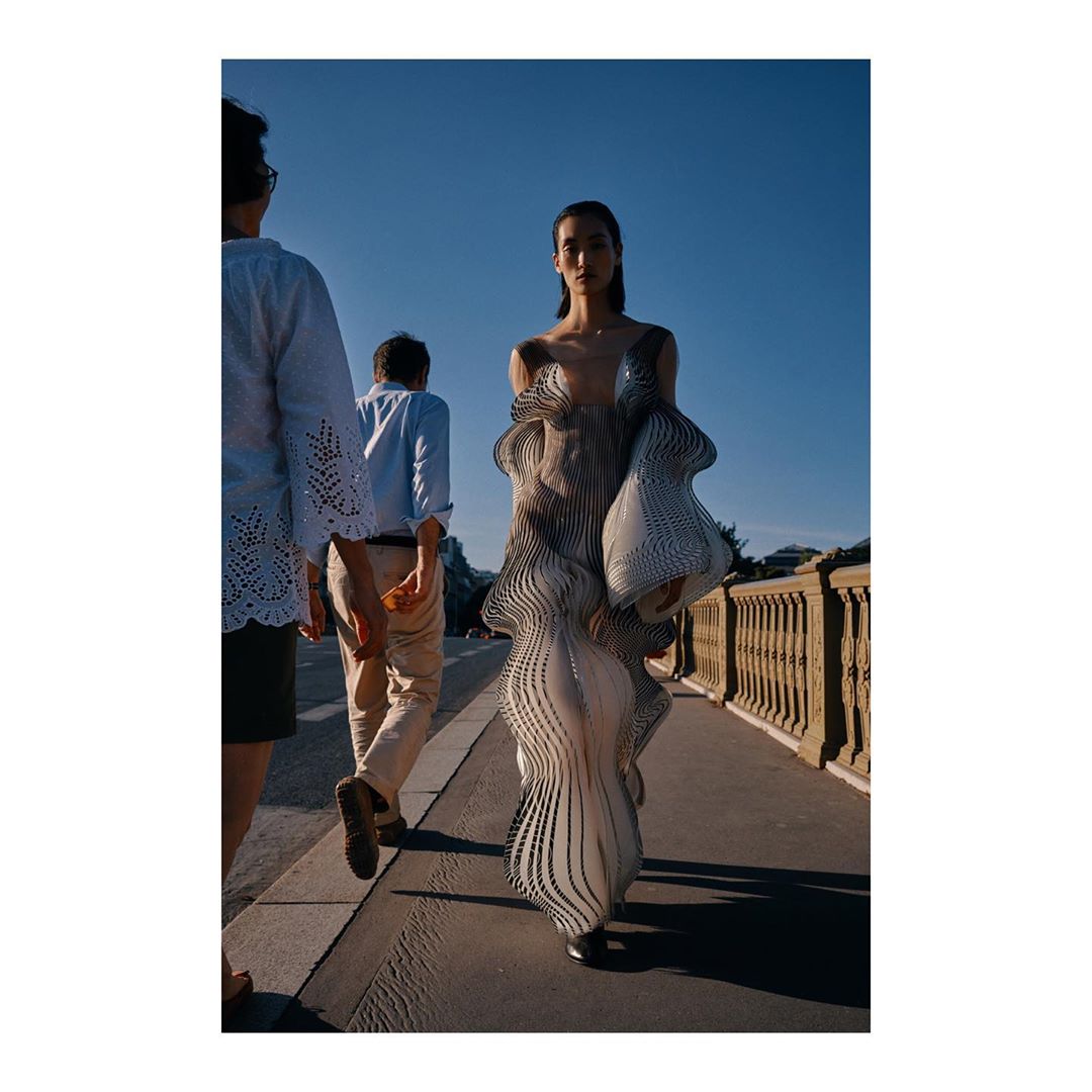 Woman in futuristic dress walking on bridge, flanked by two blurred figures against a clear blue sky in a fashion editorial shot.