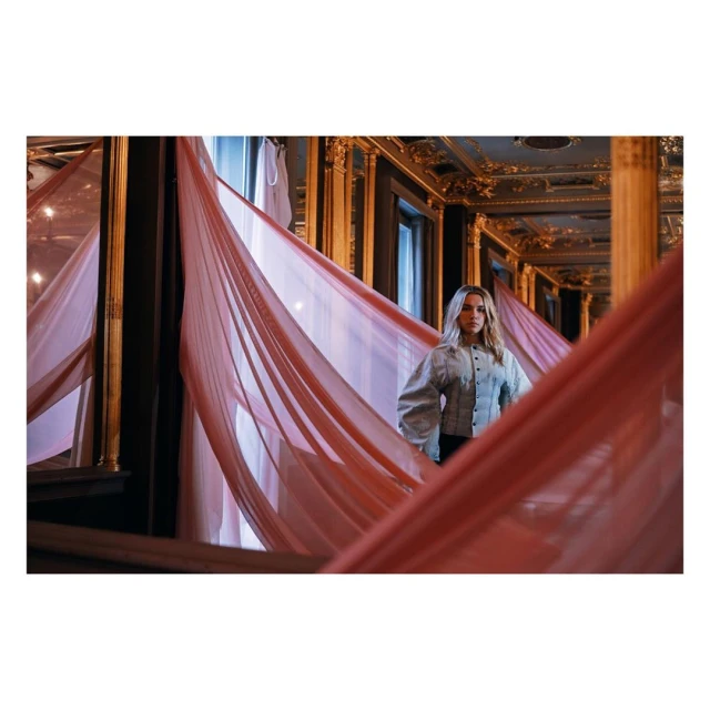 Indoor shot of a blonde adult female wearing a grey jacket, standing amidst layers of draped pink fabric in an ornate interior with mirrors.