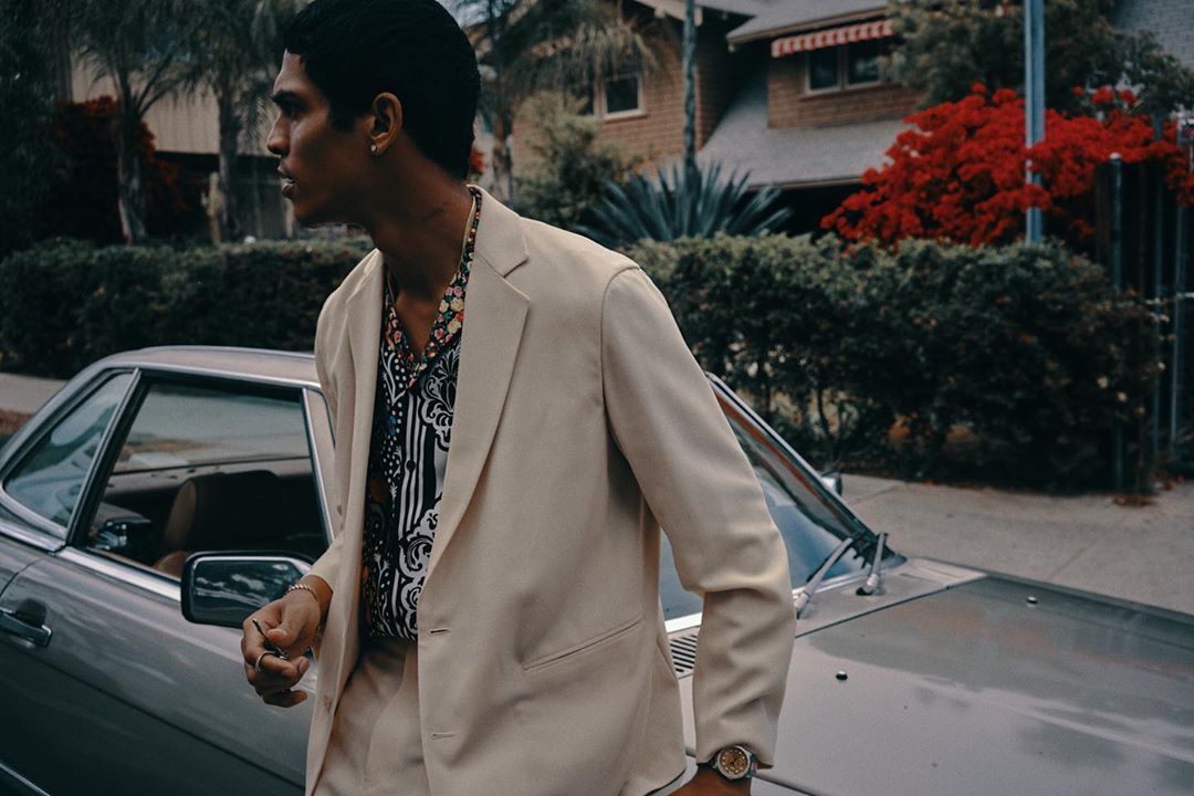 Adult male in a cream suit leaning against a silver car with a house and trees in the background; outdoor fashion shot.
