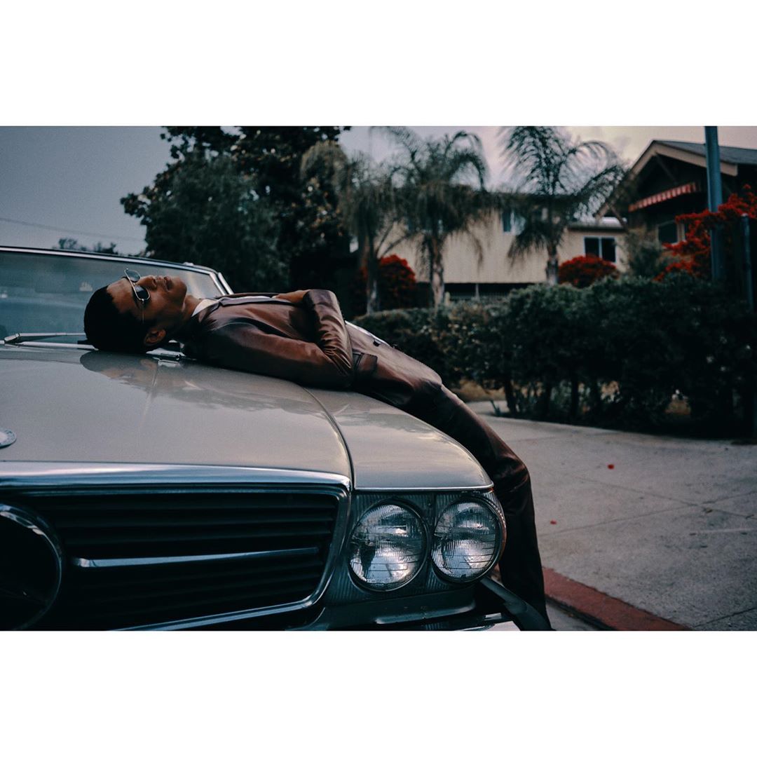 Young man in suit and glasses reclines on the hood of a convertible in front of a house on a tree-lined street.