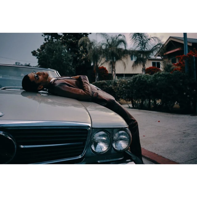 Young man in suit and glasses reclines on the hood of a convertible in front of a house on a tree-lined street.