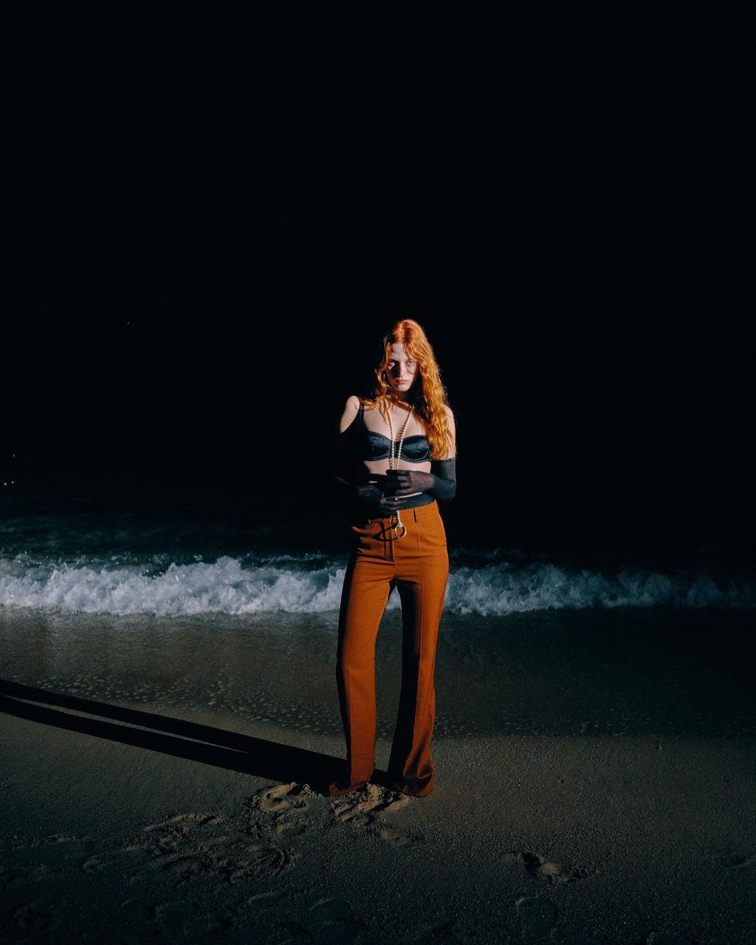 Red-haired woman standing on a sandy beach at night wearing rust trousers and dark top