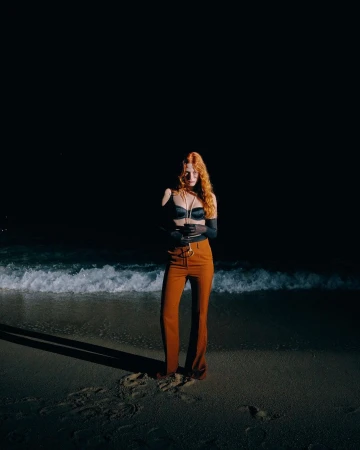 Red-haired woman standing on a sandy beach at night wearing rust trousers and dark top