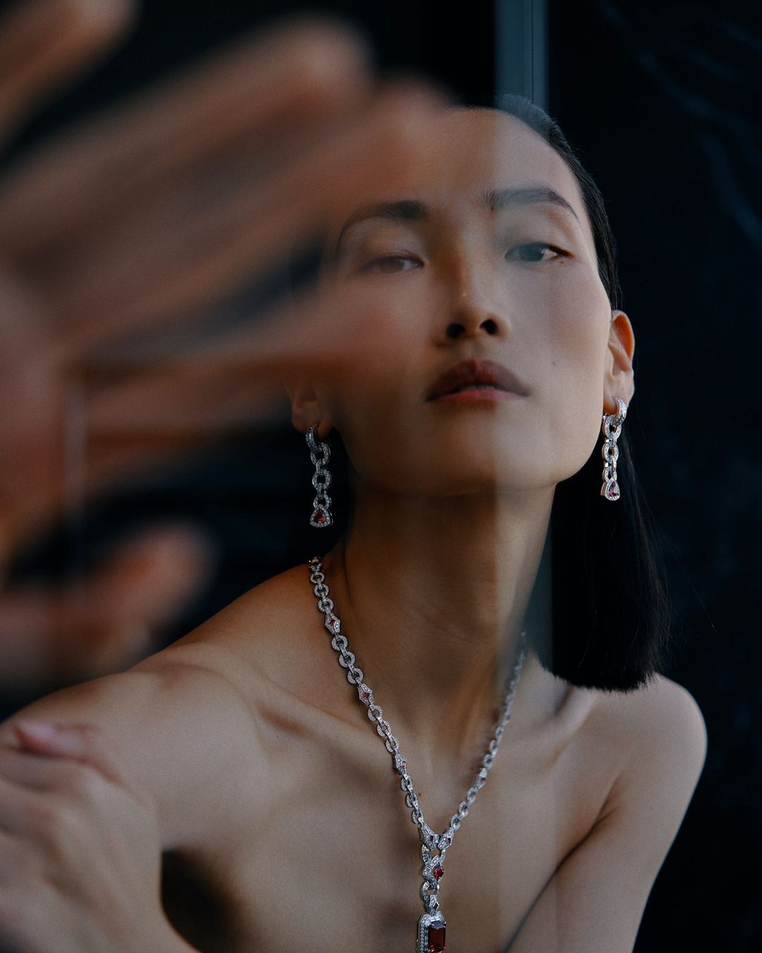 Asian woman portrait with elegant diamond jewelry necklace and earrings, close-up studio fashion shot with a hand partially obscuring her face.