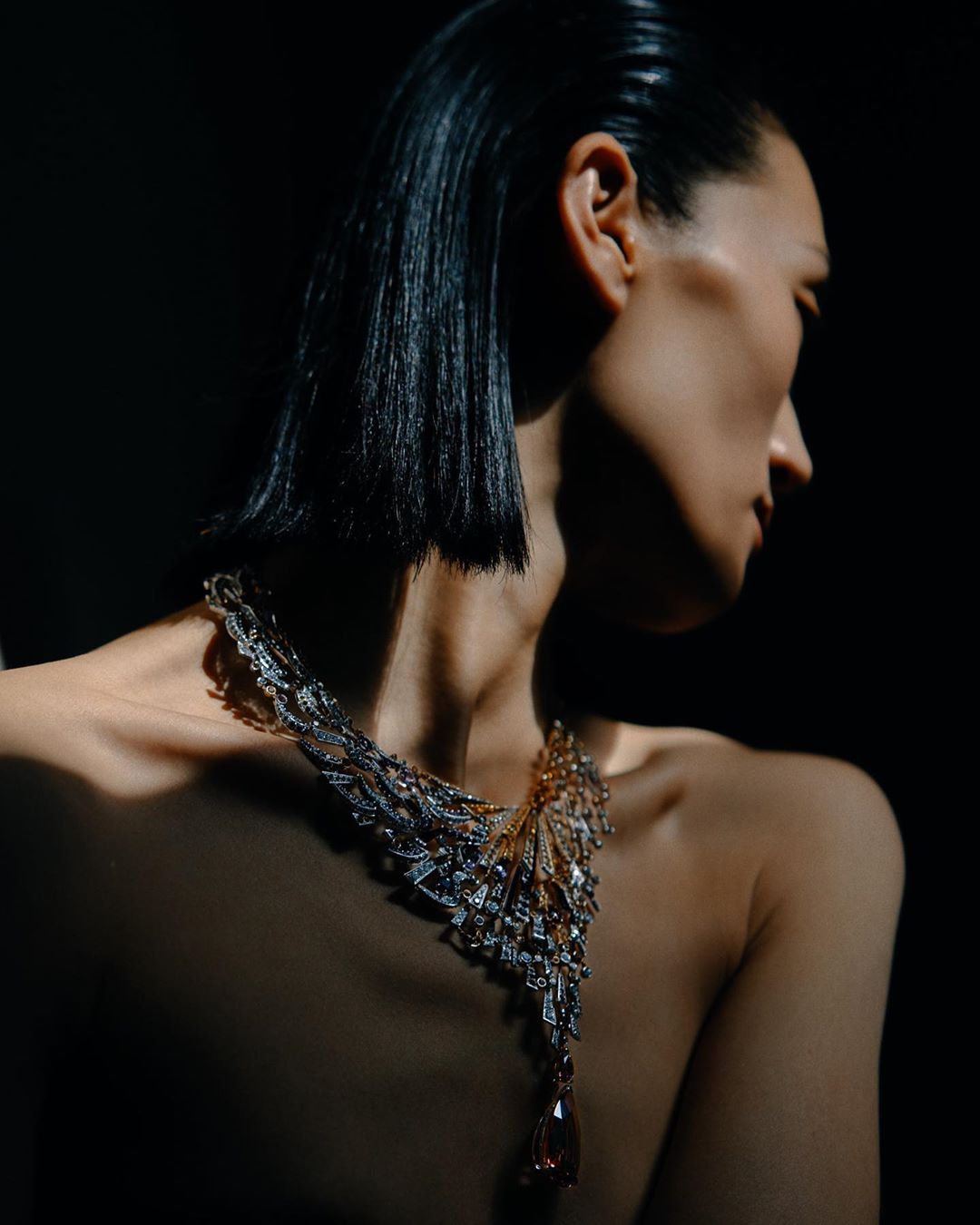 Woman with black bob hairstyle wearing a silver and gold necklace with a large orange pendant, photographed in a dark studio.