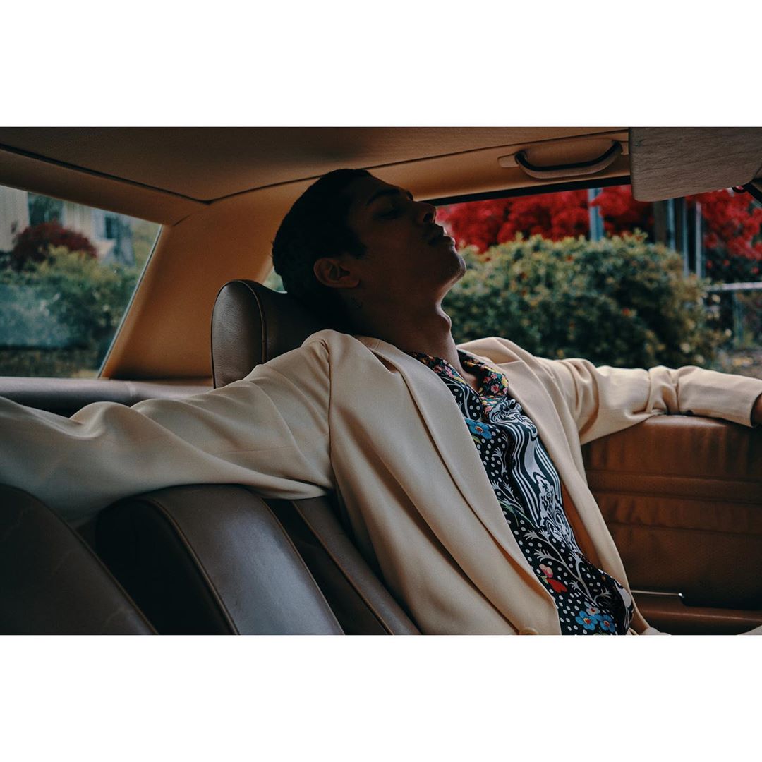Man reclines in the backseat of a car wearing a cream blazer and patterned shirt, bathed in warm light with a green outdoor backdrop.