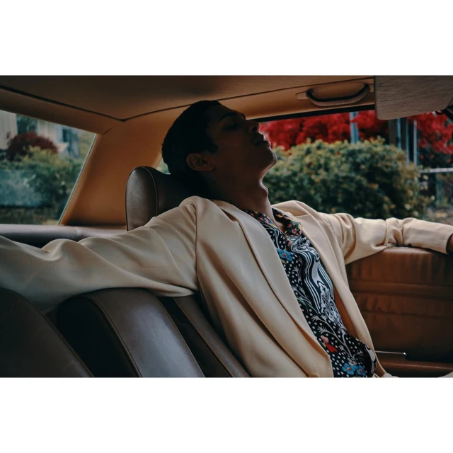 Man reclines in the backseat of a car wearing a cream blazer and patterned shirt, bathed in warm light with a green outdoor backdrop.