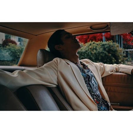 Man reclines in the backseat of a car wearing a cream blazer and patterned shirt, bathed in warm light with a green outdoor backdrop.