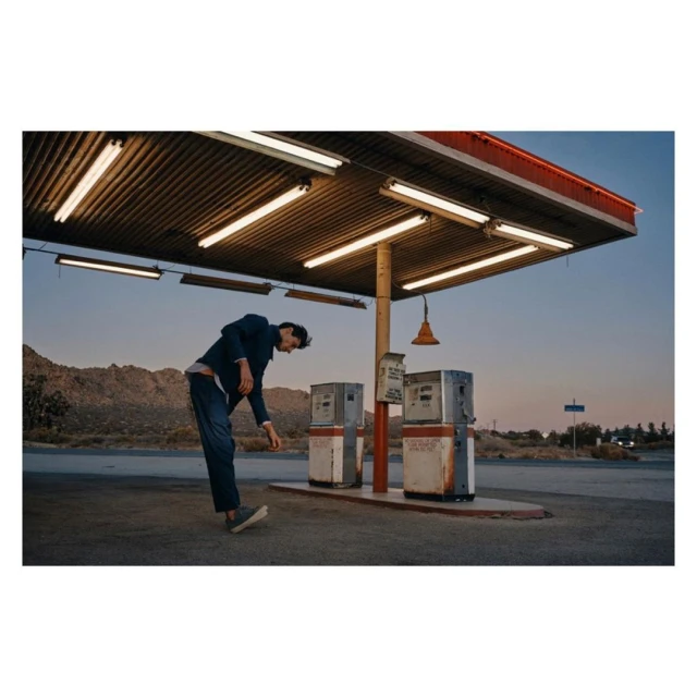 A man in a suit bends over at a vintage gas station under the awning, with mountains visible in the distance, bathed in twilight.