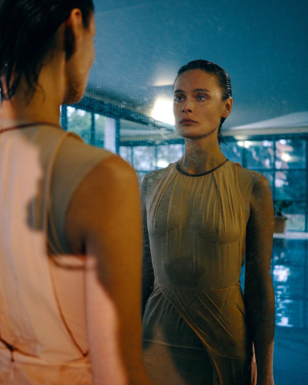 Woman in a sheer dress gazes into a mirror reflecting an indoor pool with windows to trees outside, creating a serene and reflective scene.