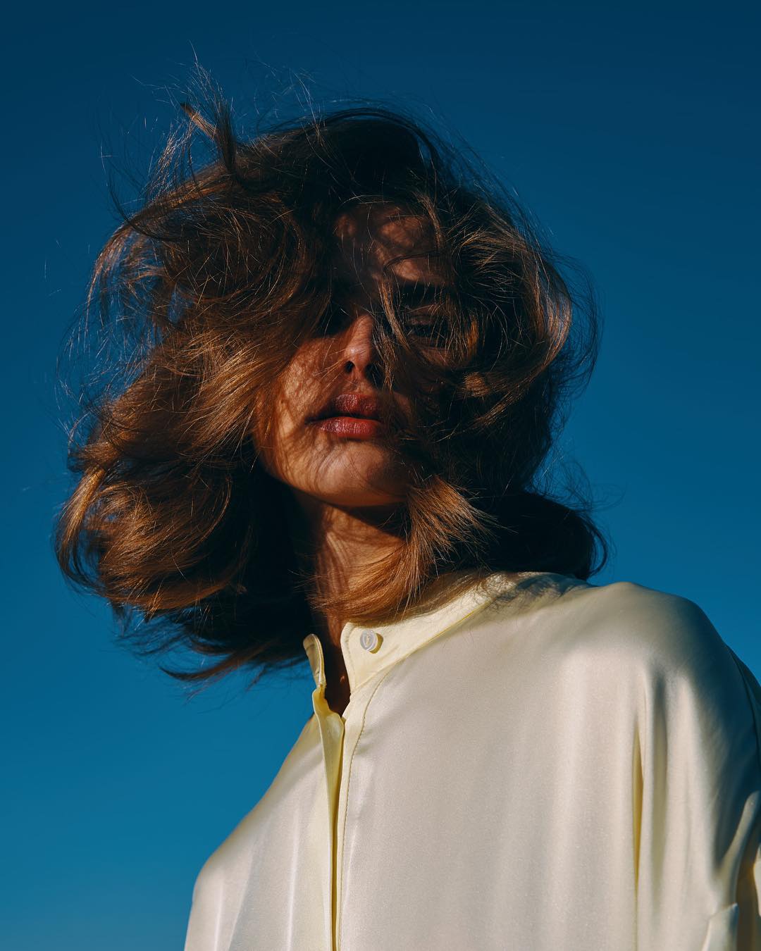 A woman with medium-length wavy blonde hair posing against a vibrant blue sky, wearing a light yellow collared shirt, in soft lighting.