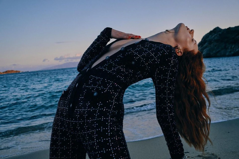 Fashion shot on a beach featuring a long-haired woman in a black and silver jumpsuit leaning backwards with the ocean as the backdrop.