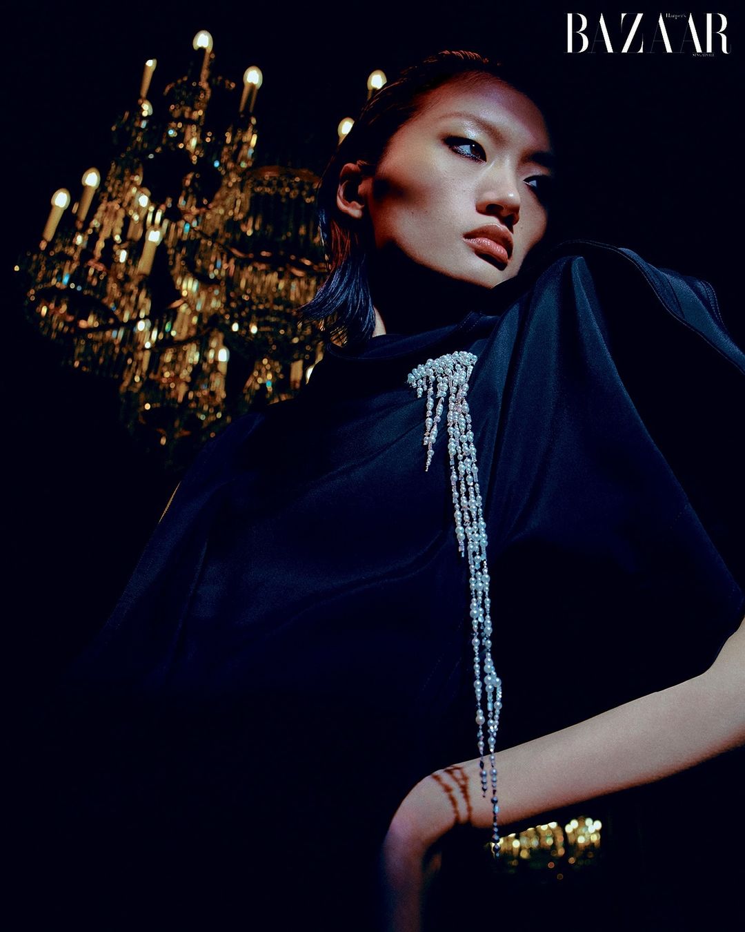 Elegant Asian woman in a black dress with a jeweled brooch poses in front of a lit chandelier for a fashion editorial.
