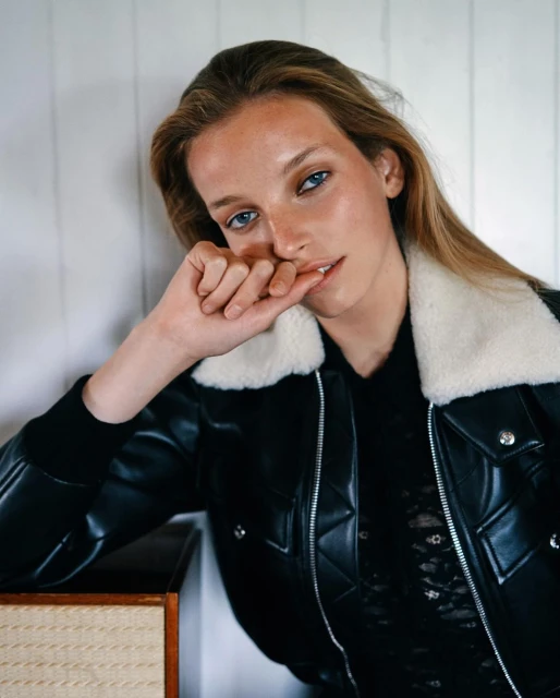 Blonde woman wearing a black leather jacket with a white fur collar in a studio setting, resting her face on her hand.