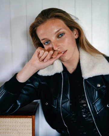 Blonde woman wearing a black leather jacket with a white fur collar in a studio setting, resting her face on her hand.
