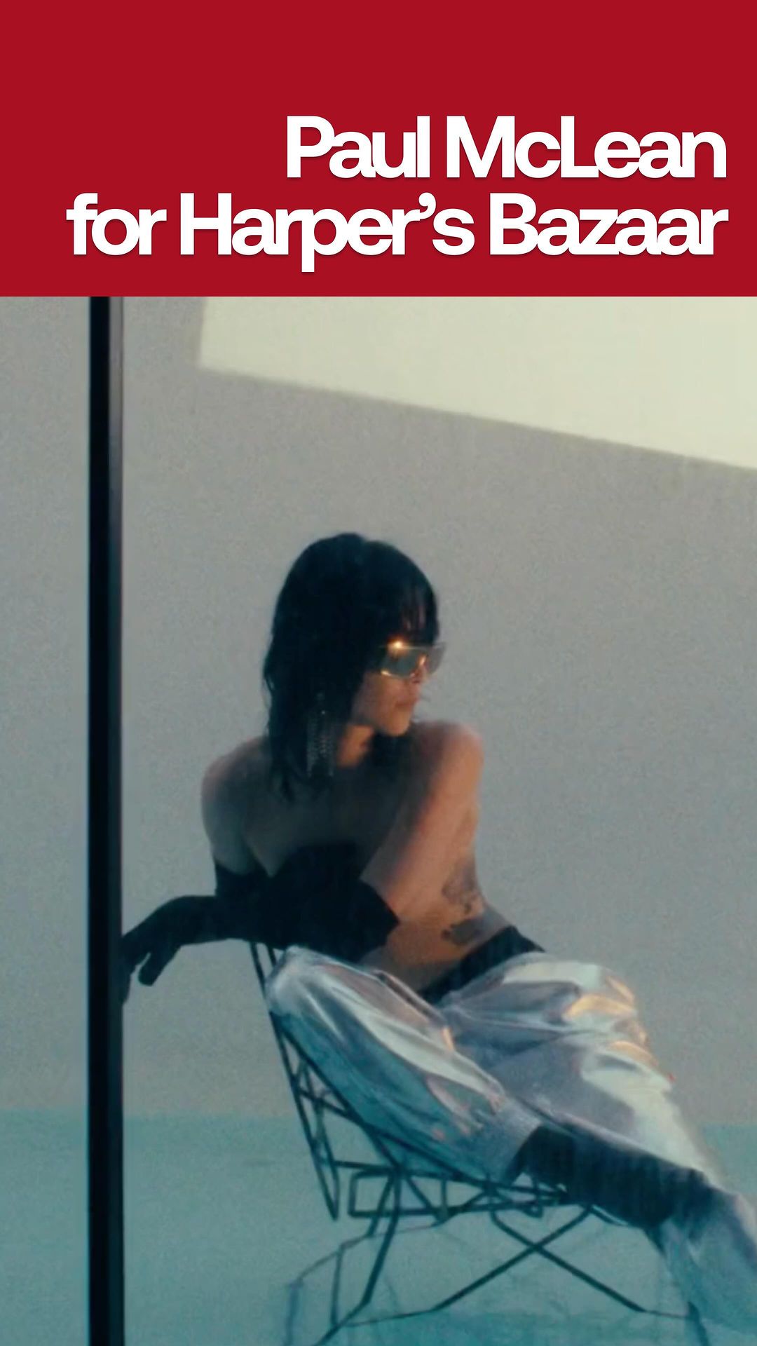 Woman with dark hair, sunglasses and metallic pants sits on wire chair in studio lit scene with blue backdrop. Fashion Editorial.