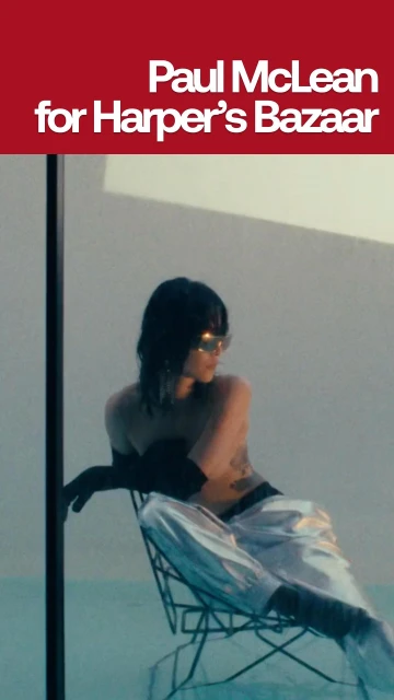 Woman with dark hair, sunglasses and metallic pants sits on wire chair in studio lit scene with blue backdrop. Fashion Editorial.