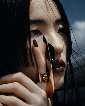 Close-up portrait of an Asian woman with dark hair and dark red lips, wearing a golden ring and a nail accessory. Sky background.