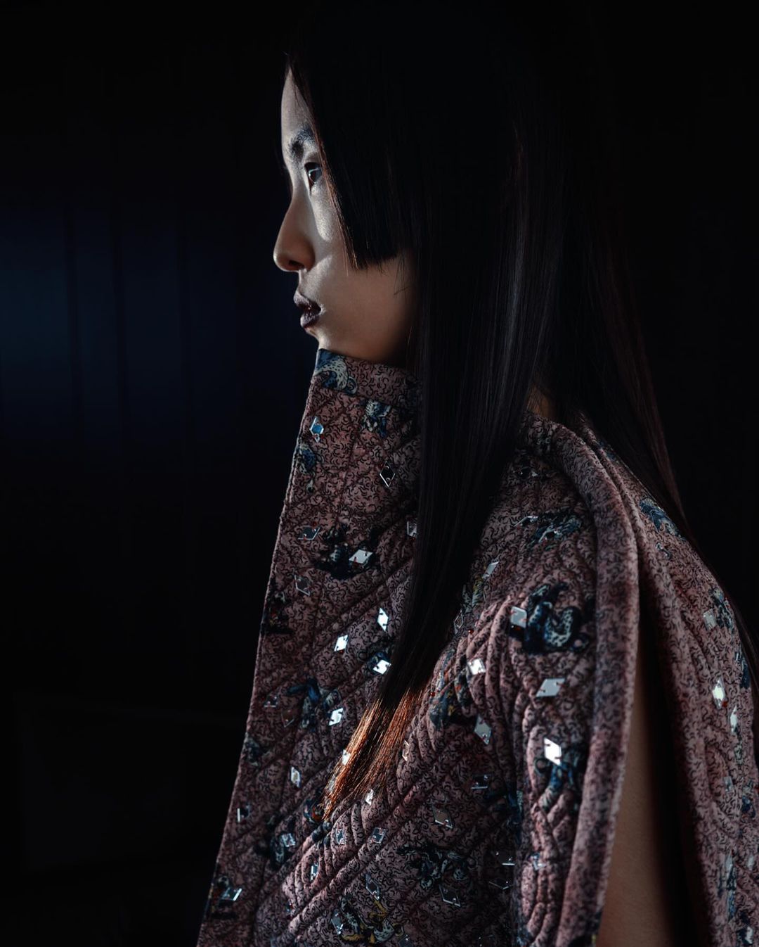 Profile portrait of an Asian female wearing a quilted, embellished cape against a dark background, showcasing fashion and style.