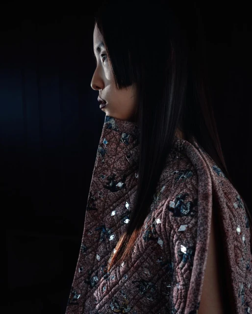 Profile portrait of an Asian female wearing a quilted, embellished cape against a dark background, showcasing fashion and style.