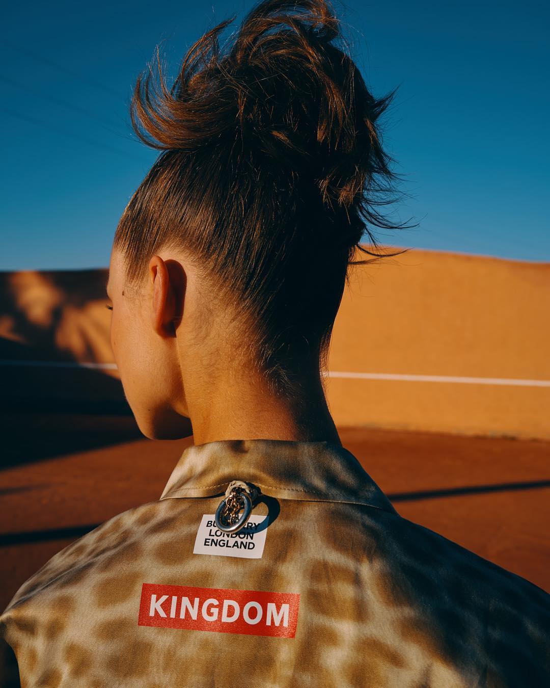 Adult female fashion model poses outdoors wearing a tan and white patterned top with 'Kingdom' text. Back view against the blue sky and sand.