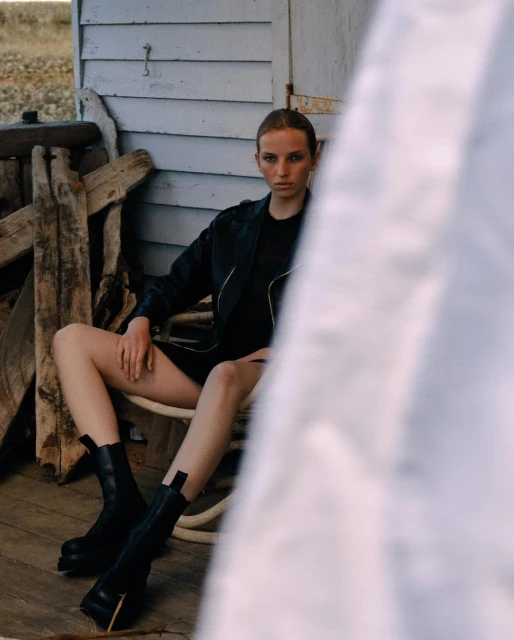 A young woman in a black dress and boots sits on a chair on a porch with weathered wood walls, posing for a fashion shot in outdoor lighting.