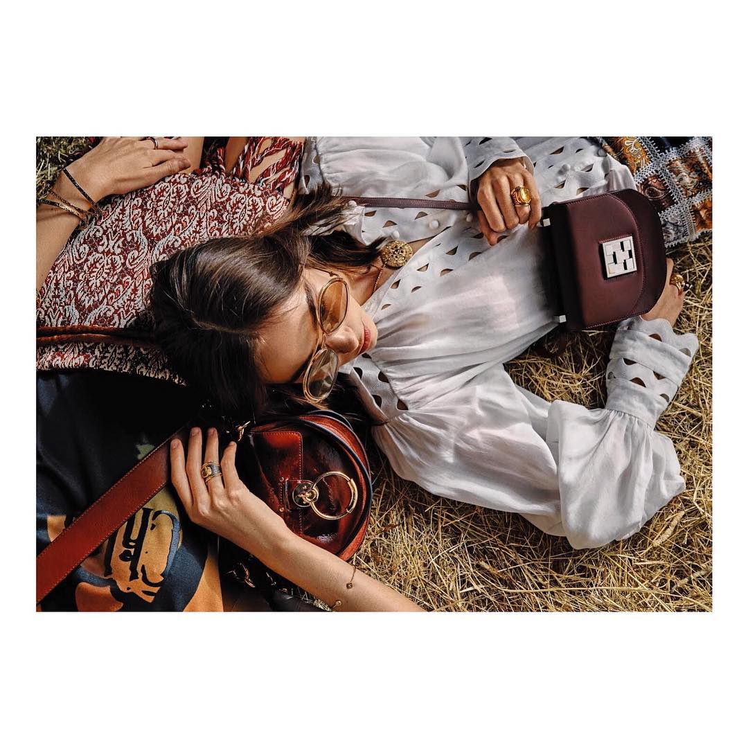 Two women lie on a blanket in an outdoor setting with brown purses and neutral-toned clothing, exuding a relaxed and fashionable vibe.