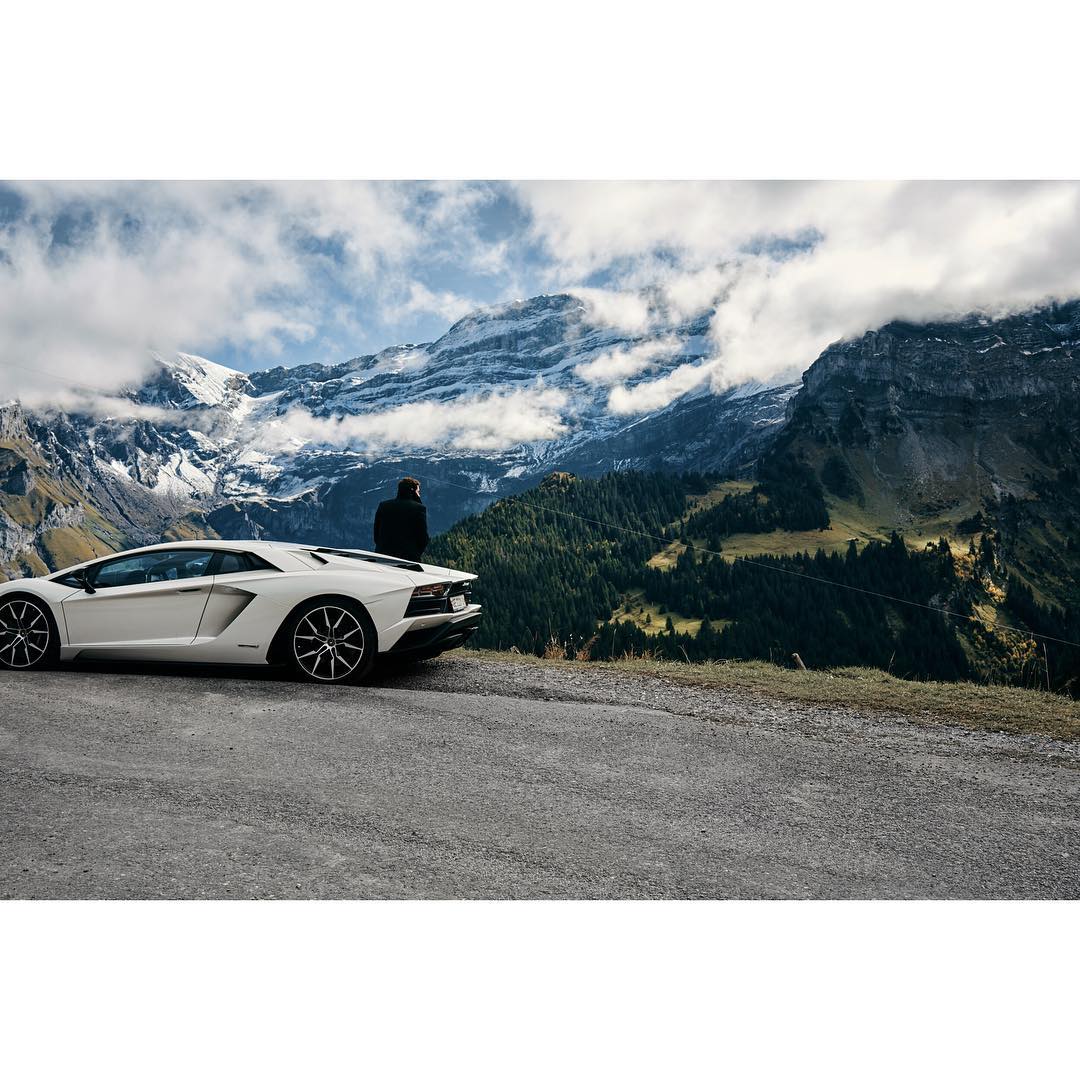 White Lamborghini sports car parked on a winding road overlooking a mountain range with a person standing next to the vehicle.