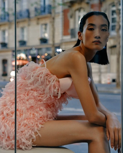 Portrait of a slender Asian woman in a textured pink dress sitting indoors near a window with a city street in the background.