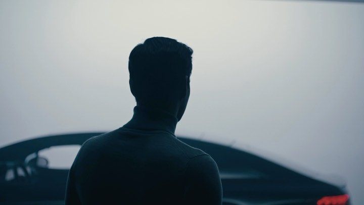 Silhouette of a man with a turtleneck sweater standing beside a black car against a gray backdrop in an indoor environment.