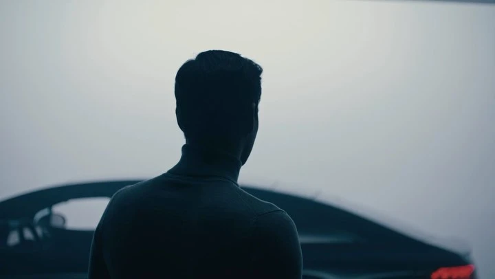 Silhouette of a man with a turtleneck sweater standing beside a black car against a gray backdrop in an indoor environment.