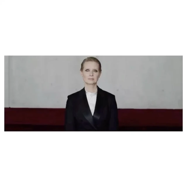 Adult woman in a dark blazer with a white top, looking at the camera with a neutral expression, in front of a two-toned wall.
