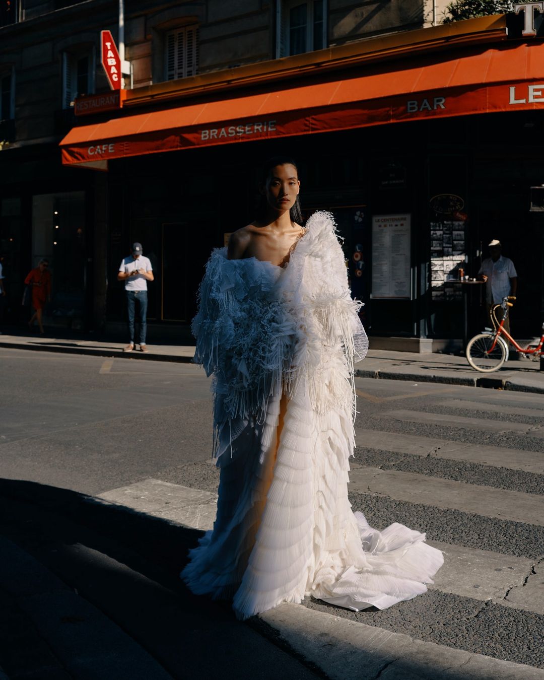 Fashion model in a large, white feathered dress stands on a city street with cafe and shops in the background, creating a striking visual.
