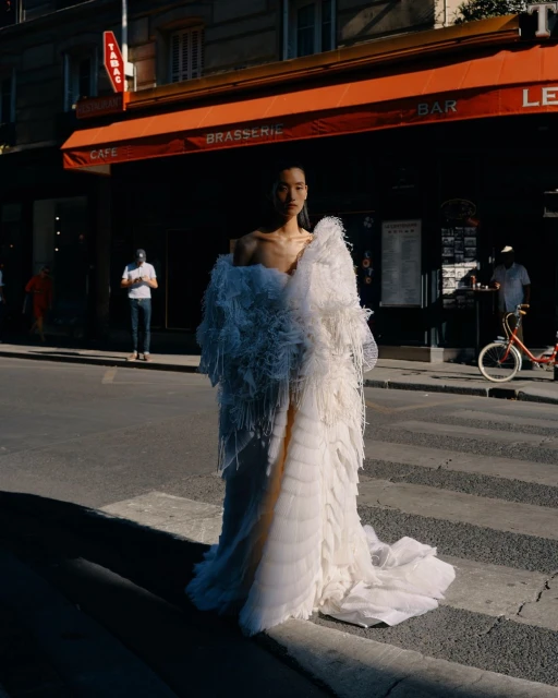 Fashion model in a large, white feathered dress stands on a city street with cafe and shops in the background, creating a striking visual.