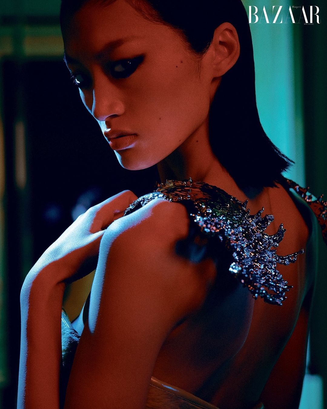 Glamorous portrait of a woman in a shimmering garment, bathed in warm, dramatic red and cool blue light, with BAZAAR logo in the upper corner.