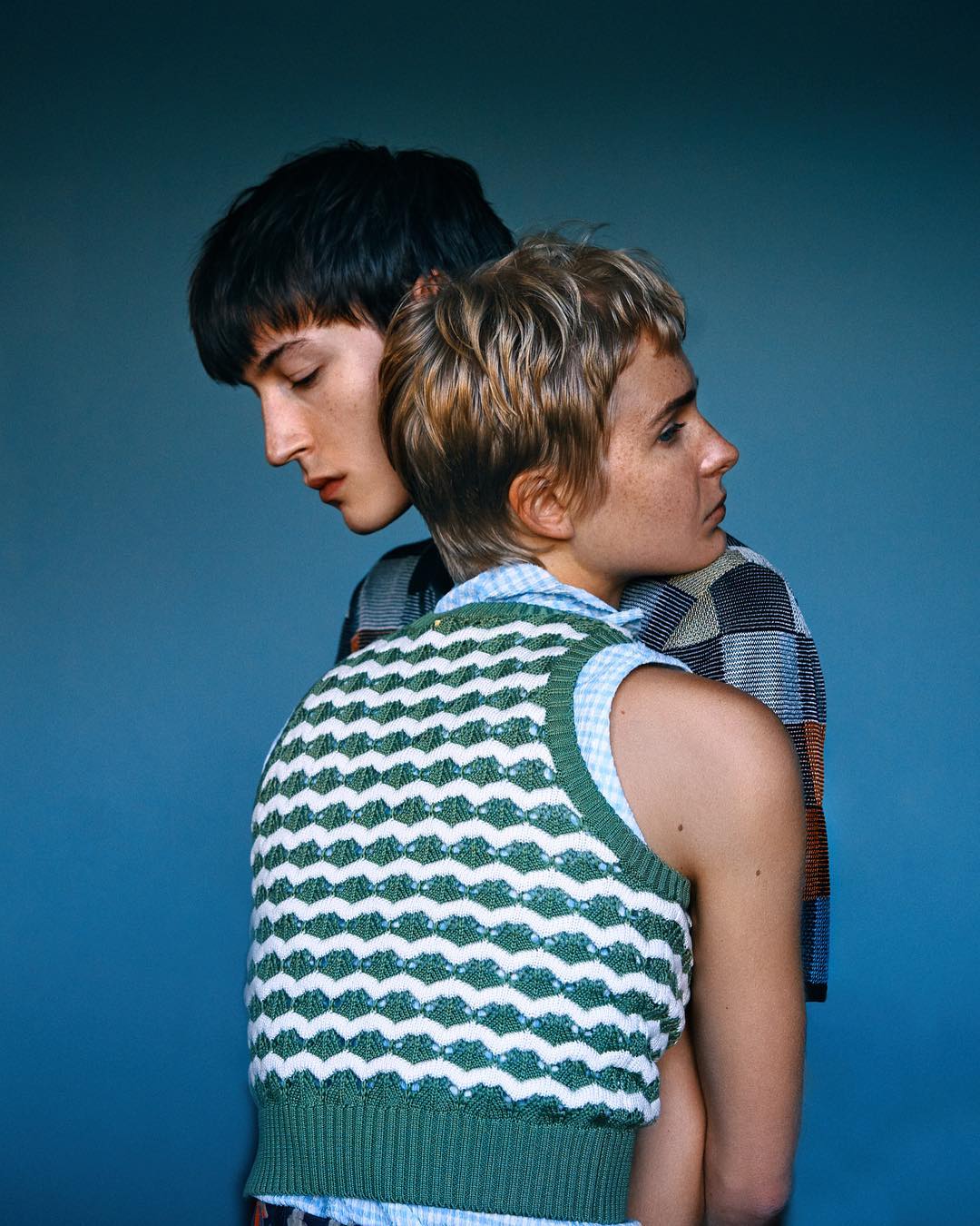 Two young people in a close portrait: a person with short blonde hair in a vest next to a person with dark hair on a blue background.