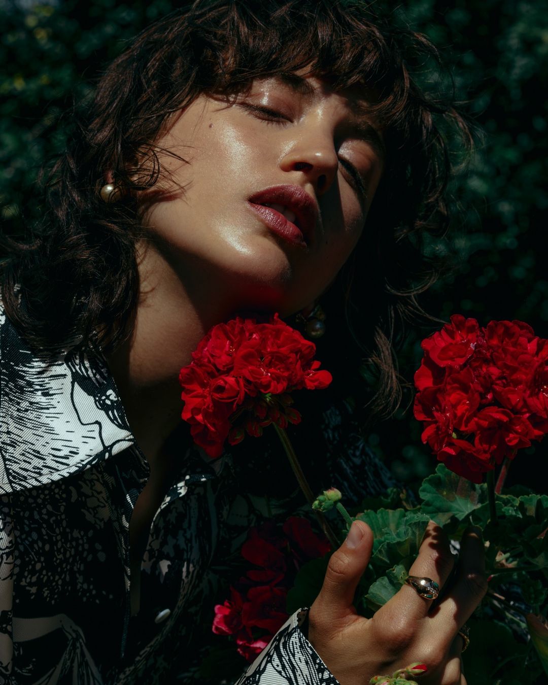 Portrait of a woman with eyes closed holding red flowers, wearing a patterned shirt, posing outdoors in a garden with natural light.
