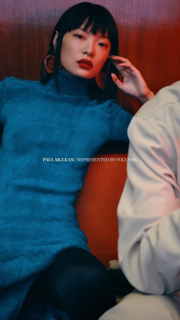 A fashionable Asian female model in a teal dress poses indoors with red lighting and wooden paneling in a close-up portrait.