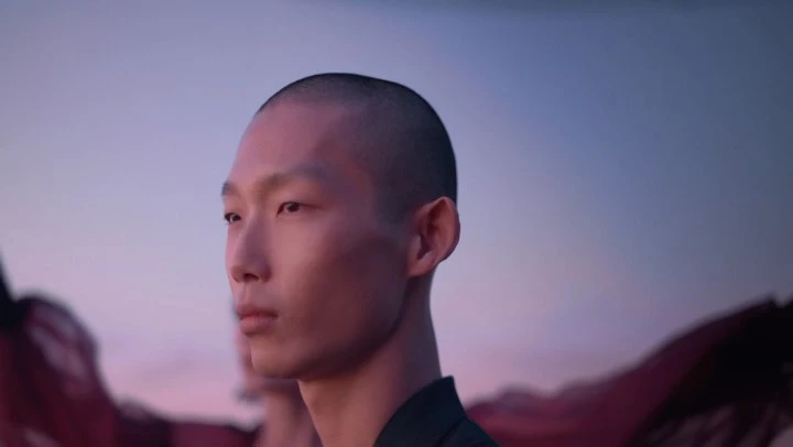 Close-up of a stylish, shaved-head person looking sideways, wearing a black jacket, and set against a background of blue and pink hues.
