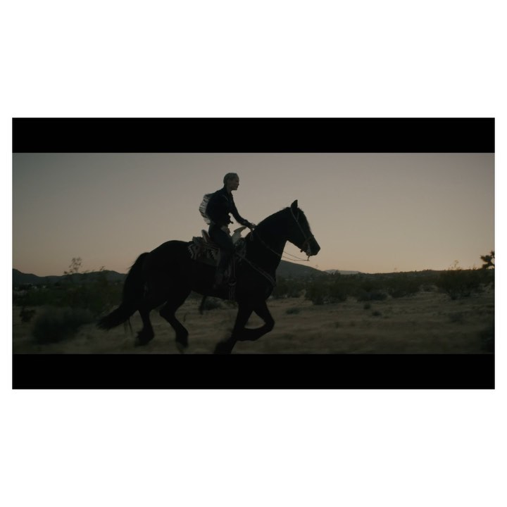 Silhouette of a person riding a dark horse through a desert landscape at dusk, with sparse vegetation and an open sky.