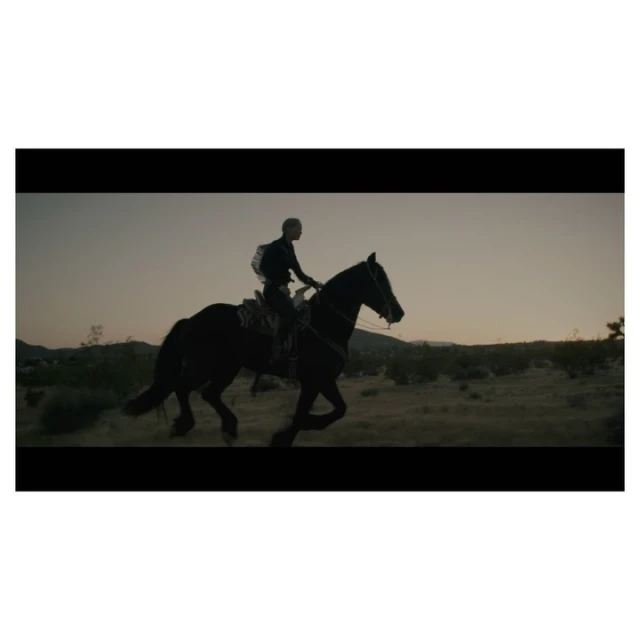 Silhouette of a person riding a dark horse through a desert landscape at dusk, with sparse vegetation and an open sky.