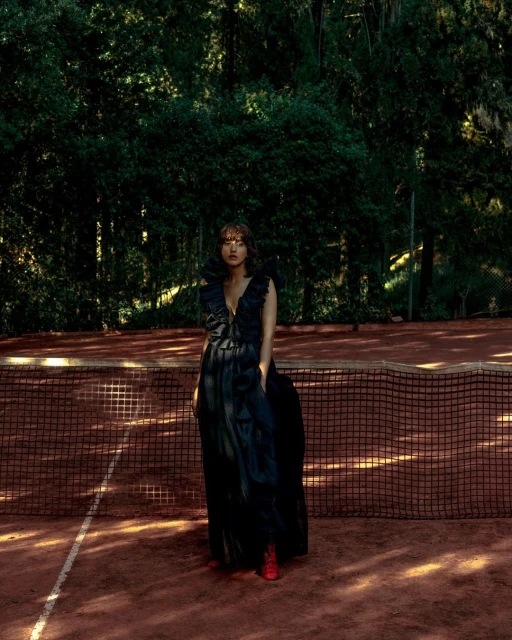 Woman in a long black dress stands on a red clay tennis court, posing in front of a net with trees in the background.