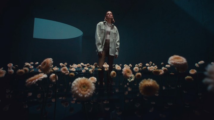 Adult woman stands in a field of daisy flowers under a dark teal background with light teal arch in a studio lighting environment.