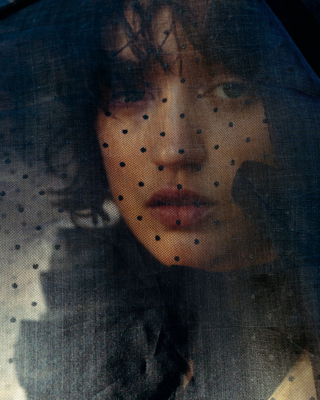 Close-up portrait of a young woman with dark hair, looking directly at the camera through a semi-transparent black polka dot veil.