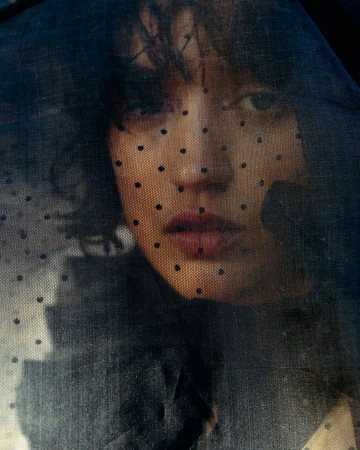 Close-up portrait of a young woman with dark hair, looking directly at the camera through a semi-transparent black polka dot veil.