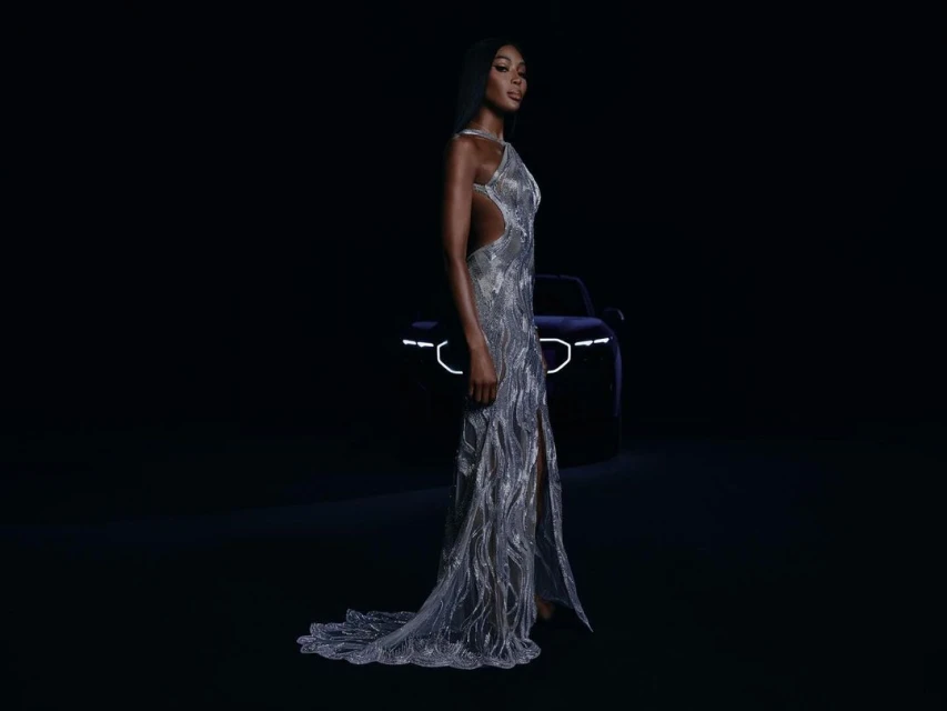 Elegant African-American woman in a glittering gown poses in front of a blurred-out luxury car with illuminated lights in a dark setting.