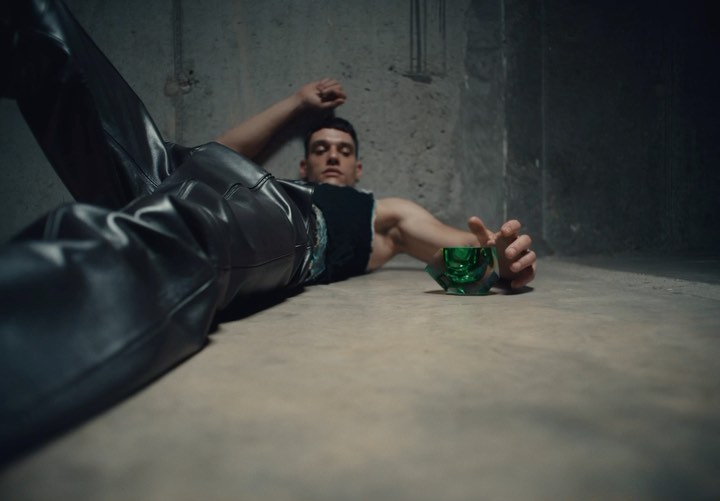 Man in leather pants reclines on floor next to green glass; against a textured grey wall in a dimly lit indoor setting.