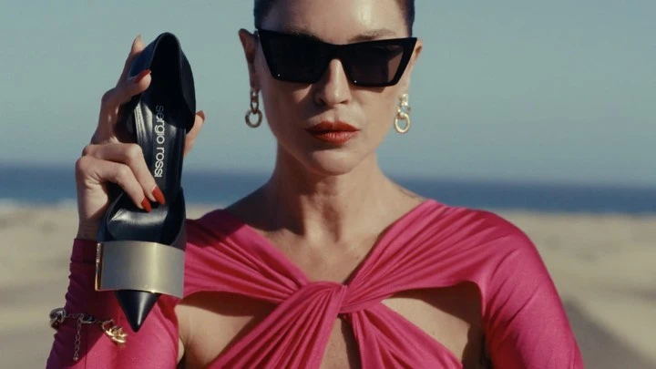 Woman in pink dress wearing sunglasses holding up a black high-heeled shoe with gold detail outdoors by the ocean